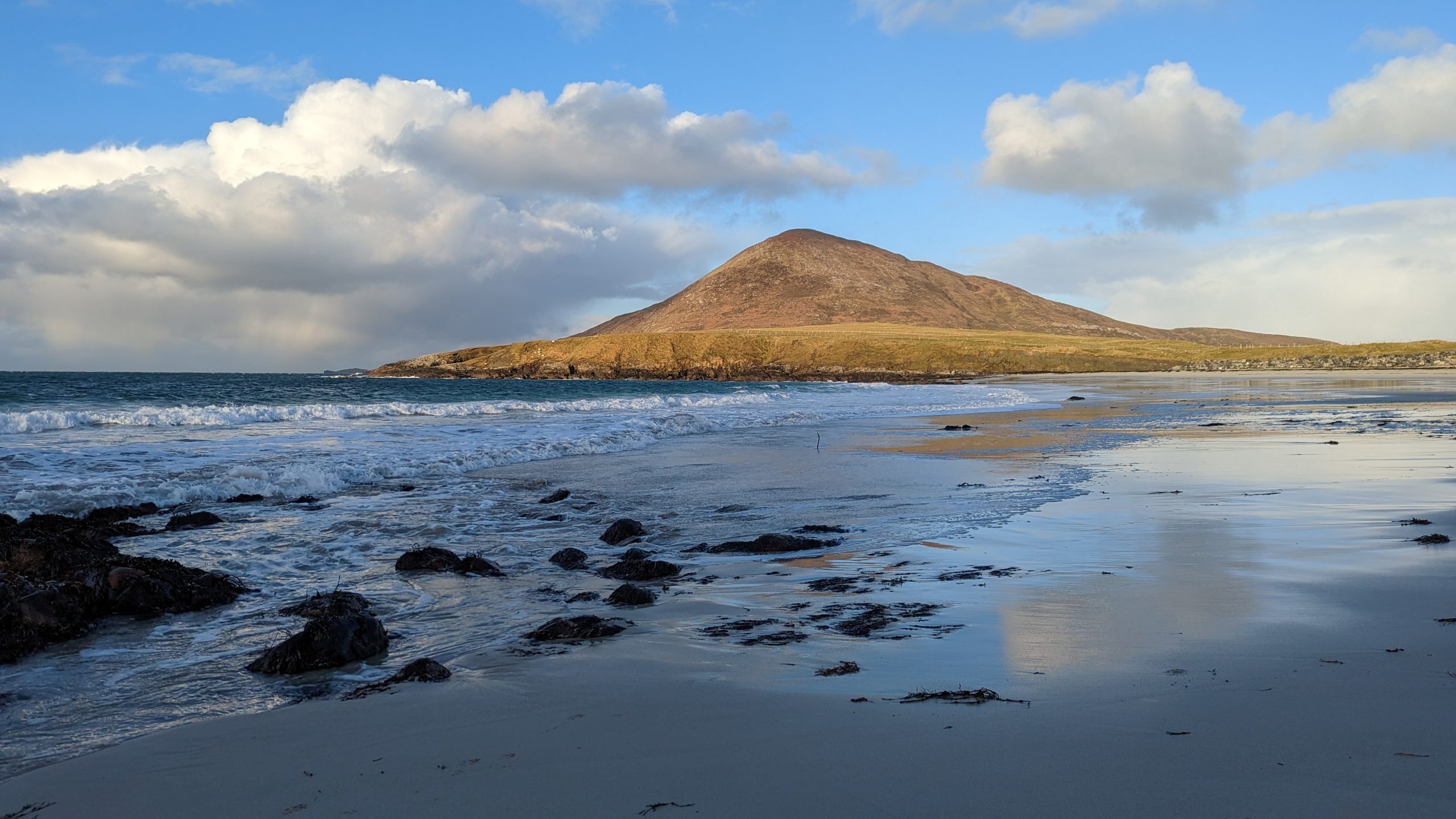 A wide sandy beach at low tide with a rounded hill across the bay